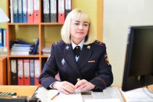 Blonde military woman at desk, monitors, office backdrop with folders, documents.