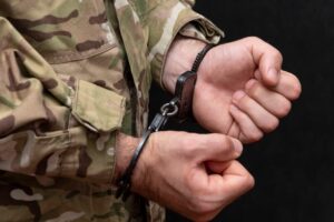 A soldier clad in camouflage attire stands restrained by handcuffs against a stark black backdrop.